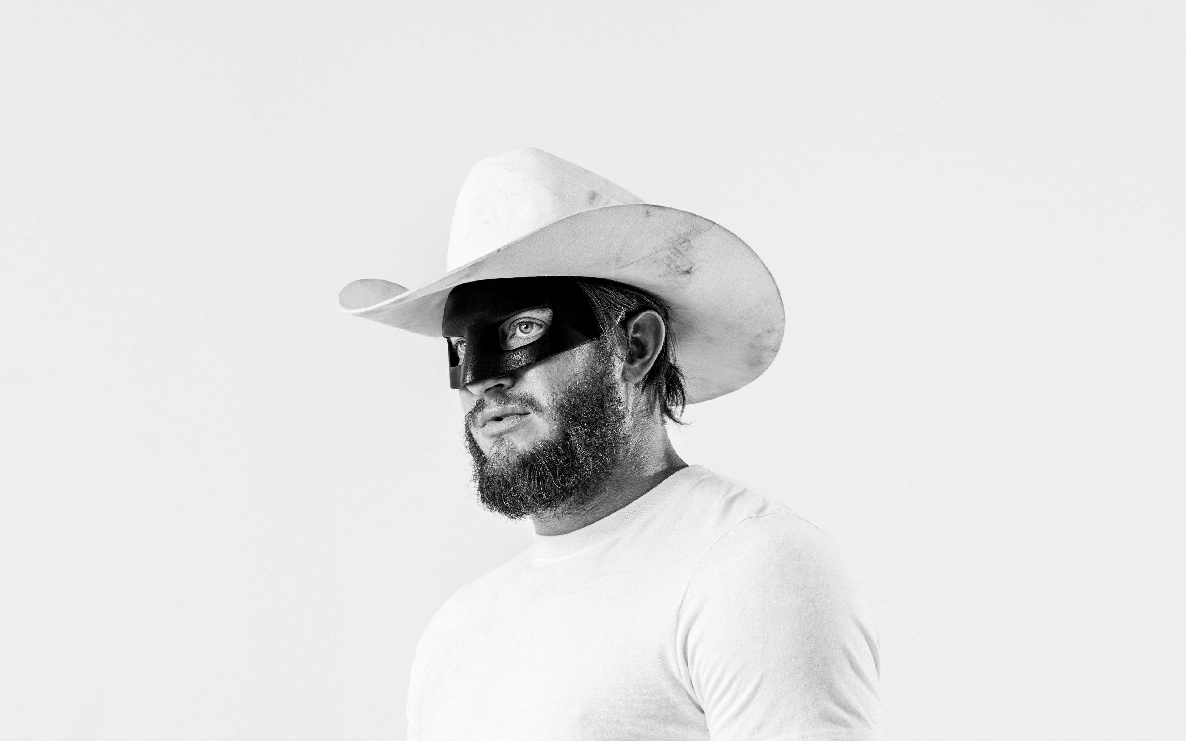 A black and white studio photograph of a Orville Peck wearing a white cowboy hat, white t-shirt, and a black eye mask against a white background.